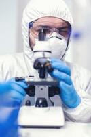 laboratorio técnico en ppe analizando virus muestra en microscopio en laboratorio. científico en protector traje sentado a lugar de trabajo utilizando moderno médico tecnología durante global epidemia. foto