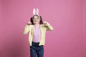 encantador niño imita un Conejo para el Pascua de Resurrección celebracion, saltando en el estudio mientras ella es vistiendo conejito orejas. alegre joven niño saltando en contra rosado fondo, interino mentecato y divertido. foto