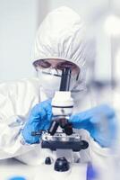 Medical engineer in ppe suit in modern laboratory looking at coronavirus sample using microscope. Chemist researcher during global pandemic with covid-19 checking sample in biochemistry lab photo