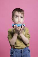 niñito jugando con un azul automóvil juguete en frente de el cámara, satisfecho niño teniendo divertido en contra rosado antecedentes. adorable niño amoroso jugar con brillantemente de colores vehículo en estudio. foto