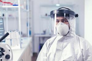 Fatigued medical scientist sitting at workplace wearing coverall with face mask against covid19 . Overworked researcher dressed in protective suit against invection with coronavirus during global epidemic. photo