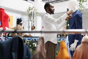 Clothing store employee adjusting male suit on mannequin while working in shopping mall. Fashion boutique african american assistant fastening formal jacket on dummy model photo