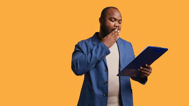 Man browsing though financial audit report on clipboard, yawning, feeling tired, studio background. Bored african american worker exhausted while reading paperwork on notepad, camera B photo