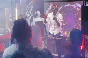 Dj playing on stage while people having fun at disco party in nightclub. Young african american man performing electronic music for clubbers crowd at discotheque in club with spotlights photo