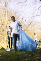 African american volunteer tidying the woods area of garbage and plastic bottles, collecting trash with claw and bags. Young woman doing voluntary work to conserve natural habitat. photo