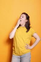 Portrait of a drowsy, young Caucasian lady in a studio, covering her mouth and yawning. Weary woman with hand on her waist, posing in front of camera and drifting off to sleep. photo