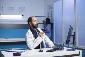 médico en un hospital comunica mediante un llamada utilizando su escritorio computadora. caucásico hombre sentado en el clínica oficina teniendo un virtual reunión. colaboración y tecnología en cuidado de la salud. foto