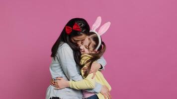 encantador Chica de escuela con conejito orejas un su mamá ondulación en cámara, teniendo divertido y riendo en contra rosado antecedentes. alegre madre y su hija abrazando y besos cada otro. cámara una. foto