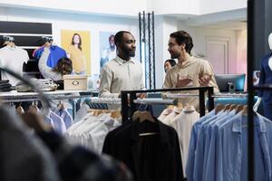 Clothing store young man worker showcasing trendy stylish apparel on rack to client. Retail market fashion boutique african american customer holding outfit hanger and talking with consultant photo