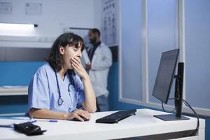 Fatigued female practitioner in modern hospital office, using digital technology for collaboration and communication. Dedicated nurse in blue scrubs tiredly reviewing medical data on desktop computer. photo