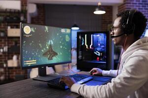 Gamer playing classic arcade action game, flying through space debris. African american man enjoying leisure time at home using high tech gaming PC to solve missions in singleplayer game photo