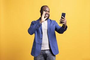 Joyful african american man greeting friends during teleconference meeting using smartphone, studio background. Happy person waving hand, saluting mate during internet call photo