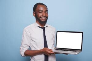 Smiling African American freelancer presents a laptop with a white screen as business mockup concept. Businessman in white shirt holds wireless computer displaying blank chromakey template. photo