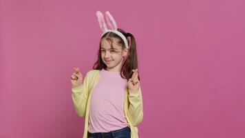 encantador dulce niña posando en estudio con dedos cruzado y esperando a recibir regalos para Pascua de Resurrección fiesta celebracion. alegre joven niño con conejito orejas preguntando para bueno suerte y fortuna. cámara una. foto