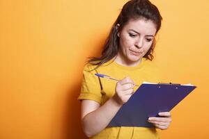 Portrait of female freelancer going through her documents and checking off items on clipboard. Caucasian lady standing in front of isolated background while using a pen to write notes. photo