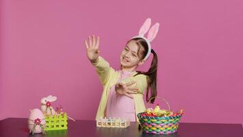 energético joven niña con adorable conejito orejas ondulación en estudio, diciendo Hola y saludo alguien mientras ella crea Pascua de Resurrección decoraciones alegre niñito posando en contra rosado fondo. cámara una. foto