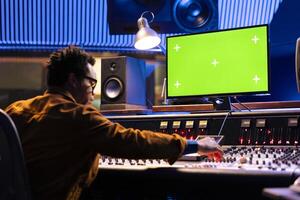 African american producer editing music on console with isolated display pc, mixing and mastering sounds on control desk board. Young audio engineer operates on sliders and buttons. photo