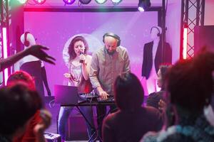 Woman singing and man dj mixing tracks while people partying on dancefloor in nightclub. People dancing and enjoying electronic music band show on stage with spotlights in club photo