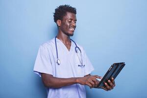 sonriente negro médico con estetoscopio es utilizando un tableta para cuidado de la salud investigación. africano americano médico profesional surf el red en su digital dispositivo, en pie en contra aislado antecedentes. foto