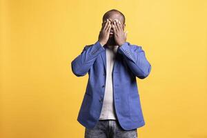 Tired BIPOC man covering face with palms, exhausted after long day at work. African american person in need of break from job, obstructing eyes with hand, isolated over studio background photo