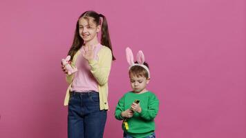 Cute brother and sister posing against pink background in studio, wearing bunny ears and playing with toys. Cheerful siblings feeling excited about easter, traditional spring holiday. Camera A. photo