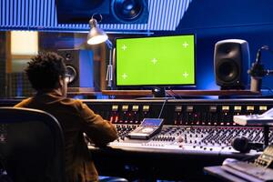 African american sound designer works with greenscreen monitor, mixing and mastering tracks on control desk panel board. Skilled audio technician twisting knobs and pushing sliders. photo