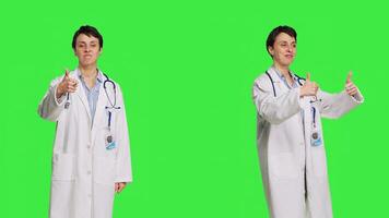 Cheerful medic doing thumbs up symbol against greenscreen backdrop, expresses positivity with like sign. General practitioner with coat giving approval and being satisfied with success. Camera B. photo