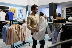 hombre participación camisa en percha, examinando estilo y adquisitivo casual vestir en Moda Al por menor almacenar. africano americano cliente comprobación formal vestir mientras elegir atuendo en boutique foto