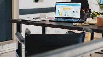 Wheelchair in office and laptop on desk showing business analytics. Medical chair in workplace offering accessibility to workers with disability and notebook with financial charts, panning shot photo