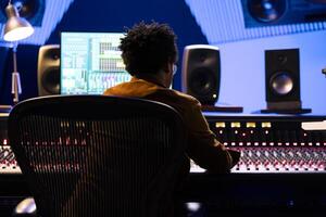 africano americano grabar ingeniero edición y mezcla canciones en estudio consola, utilizando digital software en monitor a añadir audio efectos en enviar producción. productor trabajando en nuevo música. foto