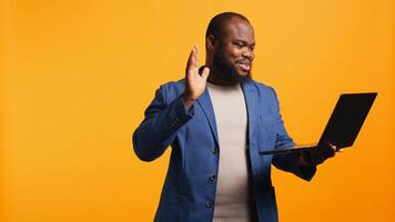 Cheerful employee greeting coworkers during teleconference meeting using laptop , studio background. Smiling worker waving hand, saluting colleagues during internet call, camera B photo