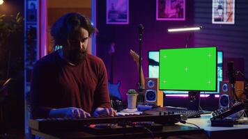 Artist learning to play at electronic piano keyboard in home studio, watching tutorial online with greenscreen display. Music producer practices playing on midi controller synthesizer. Camera A. photo