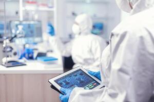 Medical engineer developing vaccine for coronavirus using tablet pc wearing ppe suit. Team of scientists conducting vaccine development using high tech technology for researching treatment against covid19 photo