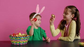 dulce niños golpes huevos juntos para Pascua de Resurrección tradicion en estudio, jugando un estacional fiesta juego en contra rosado antecedentes. encantador adorable niños teniendo divertido con festivo decoraciones cámara b. foto