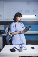 Female medical worker looking at tablet in an office while standing next to desk having pens, a clipboard, and a mouse. In the background sits a male physician working on healthcare treatments. photo