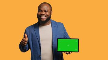 Jolly man holding isolated screen tablet, doing recommendation. Cheerful BIPOC person pointing towards chroma key device, doing thumbs up sign, studio background, camera A photo