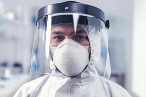 Tired healthcare scientist loooking at camera wearing ppe suit with face shieldin lab. Overworked researcher dressed in protective suit against invection with coronavirus during global epidemic. photo