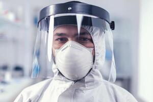 cansado cuidado de la salud científico mirando a cámara vistiendo ppe traje con cara escudo laboratorio. sobrecargado de trabajo investigador vestido en protector traje en contra invección con coronavirus durante global epidemia. foto