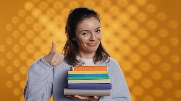 retrato de contento mujer con apilar de libros en manos demostración pulgares arriba, estudio antecedentes. jubiloso ratón de biblioteca participación pila de novelas, sensación optimista, haciendo positivo mano gesticulando, cámara si foto