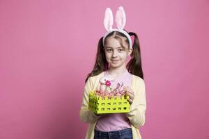 Young optimistic girl showing handmade basket decorated with eggs and a rabbit for easter festivity, posing over pink background. Little cute child with bunny ears presenting spring ornaments. photo