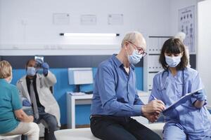 Medical specialist with clipboard and hospital document for senior male patient to sign. Nurse and elderly guy sat on clinic bed, both wearing face masks, looking at clinic insurance paperwork. photo