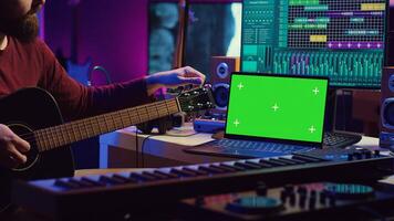 Musician learning to play new acoustic instrument in his home studio, practicing online guitar lessons on isolated greenscreen display. Watching tutorial on laptop to develop his skills. Camera A. photo