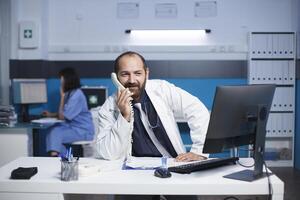 Caucasian male doctor seated at the office desk talking on the telephone with other healthcare specialists. Man wearing a lab coat is using a landline phone to speak with a hospital receptionist. photo