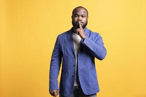 Annoyed african american man doing shushing hand gesturing, irritated by noise, having negative mood. Person placing finger on lips, doing quiet sign gesture, studio background photo