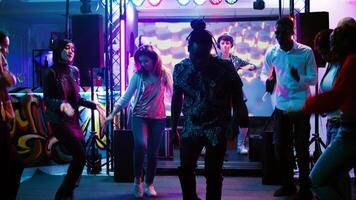 Happy persons jumping at dance party, having fun at social gathering in discotheque. Young funky friends showing dance moves on music at club, colorful spotlights. Handheld shot. photo