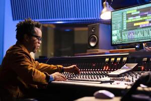 African american sound engineer works on mixing console with sliders and knobs, looking at digital software to record and process tunes. Audio technician creating new music, control room board. photo