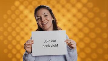 Cheerful book club president holds message urging people to join them, talking about importance of lecture, studio background. Smiling woman inviting bookworms to enlist in her association, camera B photo