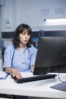 At the clinic office desk, a determined Caucasian nurse checks and analyzes her notes. Close-up shot of physician in blue scrubs getting ready to see people for medical consultations. photo