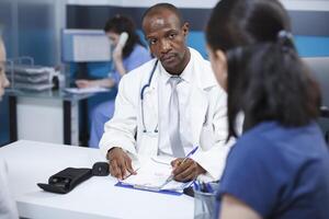 Black man wearing a lab coat, taking notes during a consultation with a female patient at the clinic. African American doctor explains the treatment plan, ensuring proper care. photo