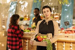 alegre hembra vendedor de frutas y verduras mercado estar propietario demostración Fresco orgánico Produce caja y mirando a cámara. ypung granjero de venta sano local Produce desde borde del camino pararse, eco productos foto